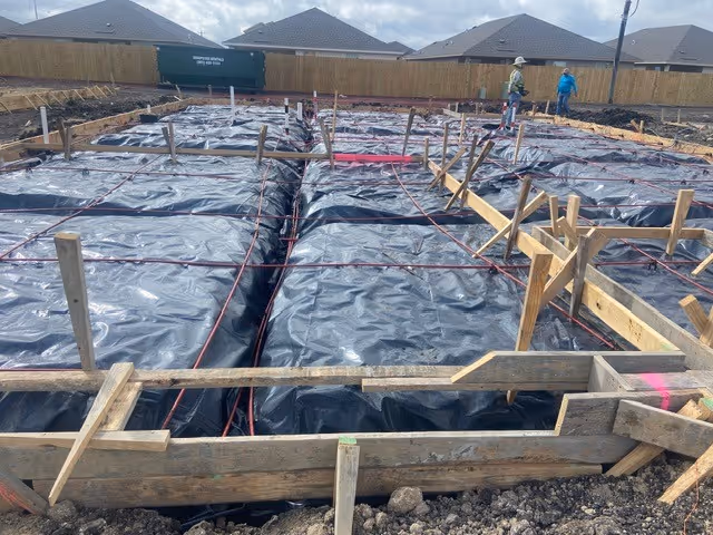Construction site with foundation trenches covered in black plastic and wooden framing, workers in background.