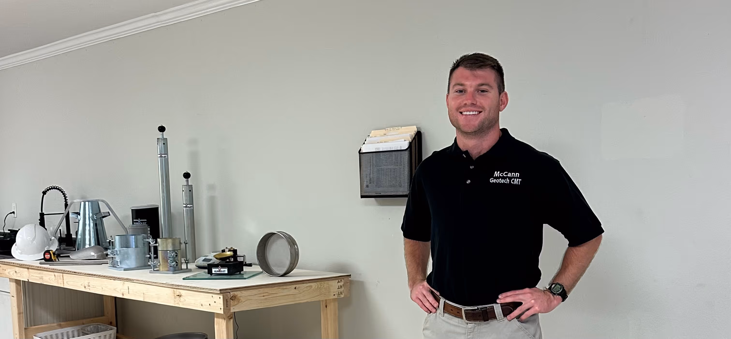 Man in black McCann Geotech CMT shirt stands smiling with hands on hips near table with testing equipment.