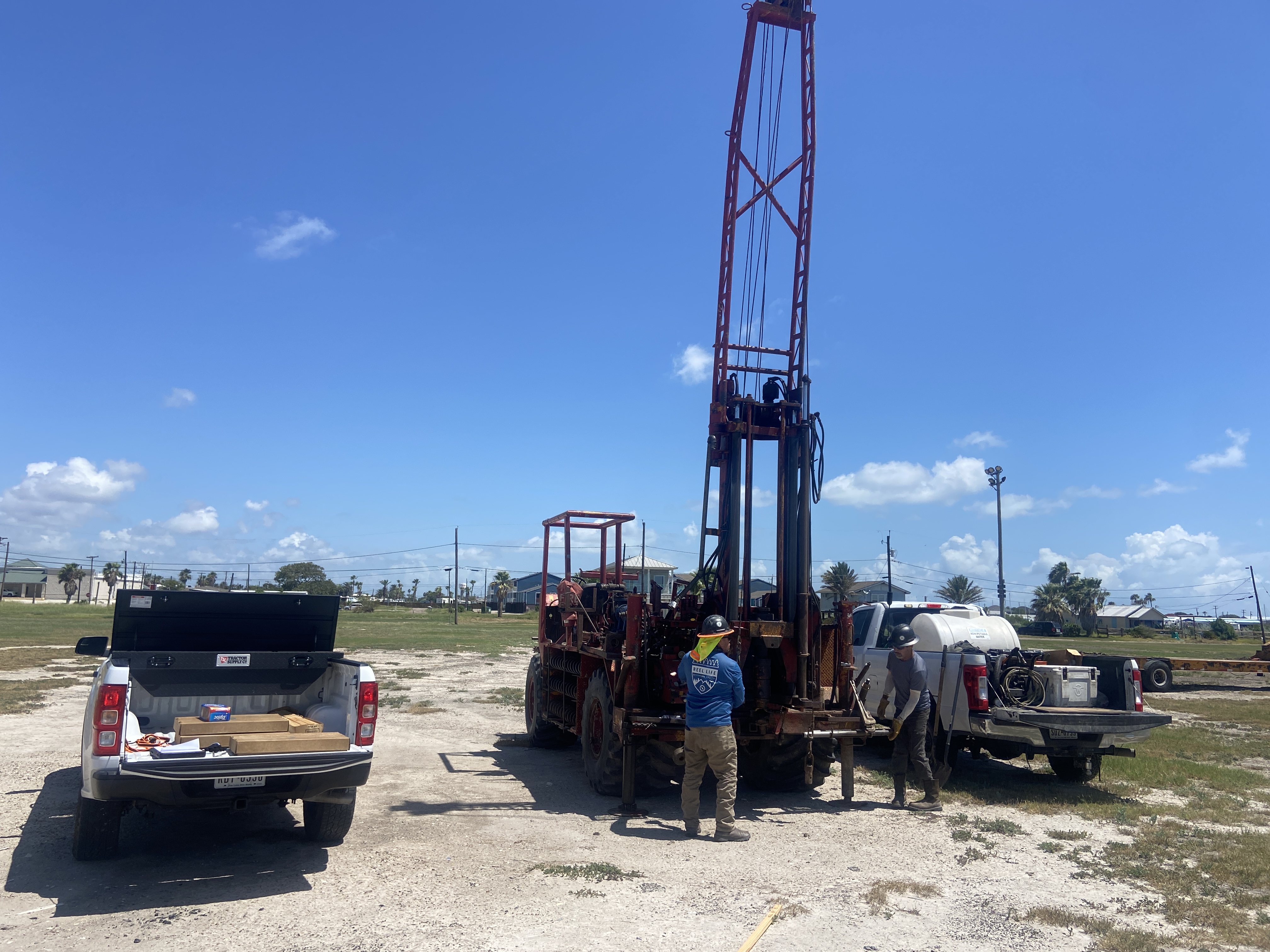 Two workers operating a red drilling rig between two white pickup trucks on a sunny day.